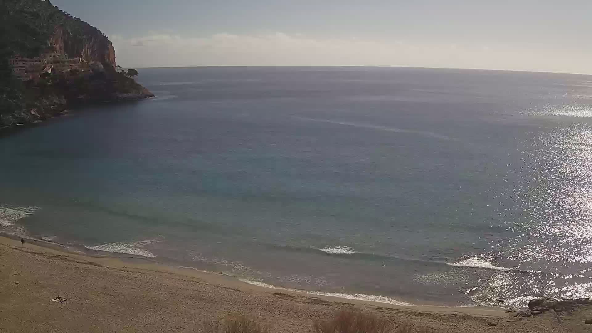 A sandy beach curves into a calm, sparkling sea next to a rocky cliff face under a mostly clear sky.