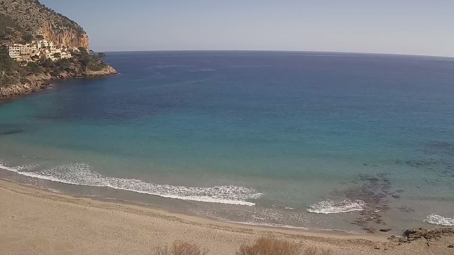 A sandy beach with gentle waves laps the shore of a clear, turquoise ocean, backed by a rocky cliff with buildings on top under a bright, sunny sky.