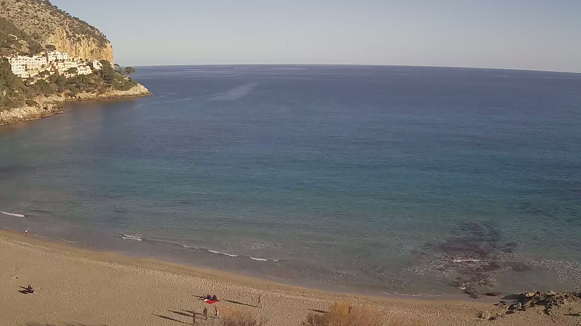 A sandy beach curves around a calm, clear ocean meeting a rocky cliff with white buildings perched on its slopes under a clear sky.