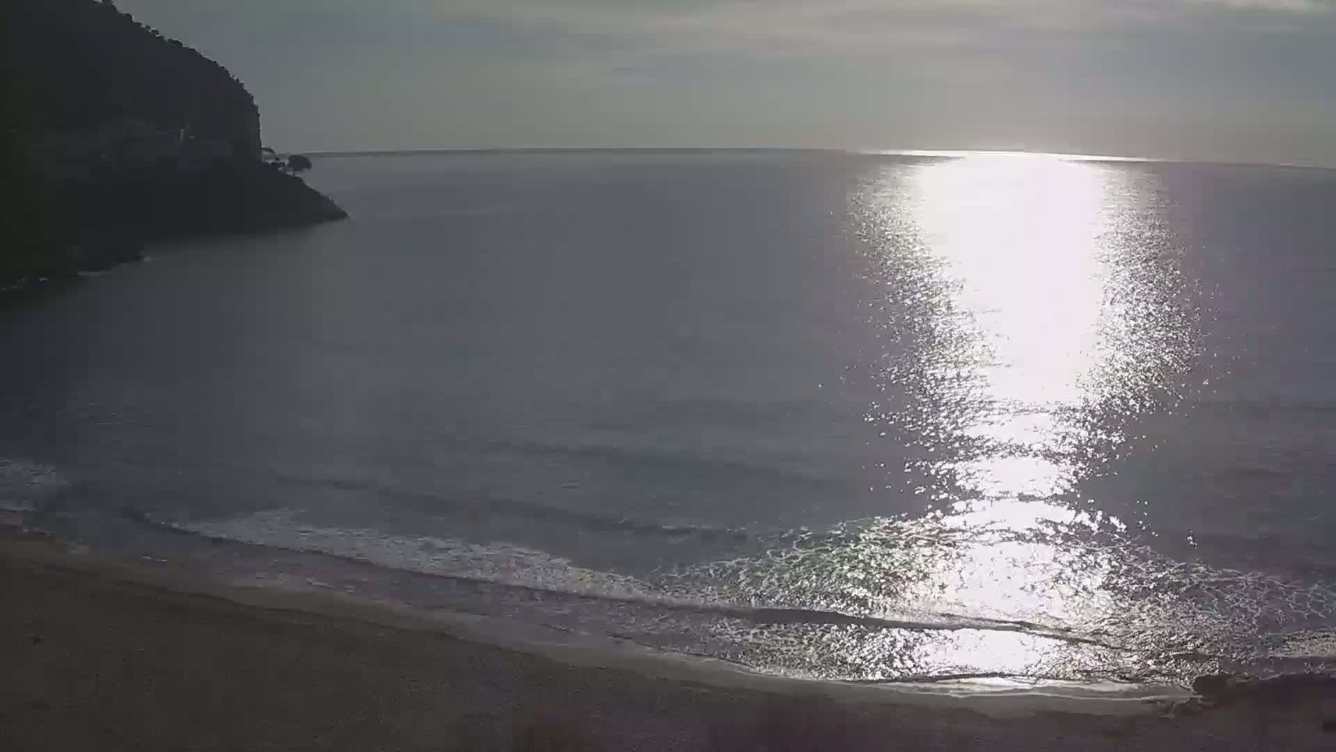 A sandy beach meets a calm sea under a bright, mostly sunny sky, with a dark cliff visible in the background.