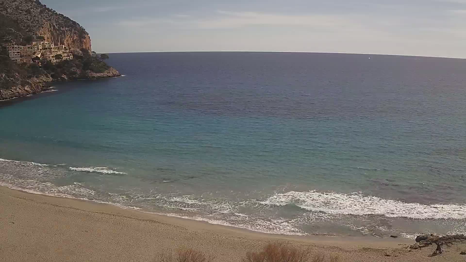 A sandy beach with gentle waves laps against the shore, under a clear sky, next to a rocky cliff with buildings on top.