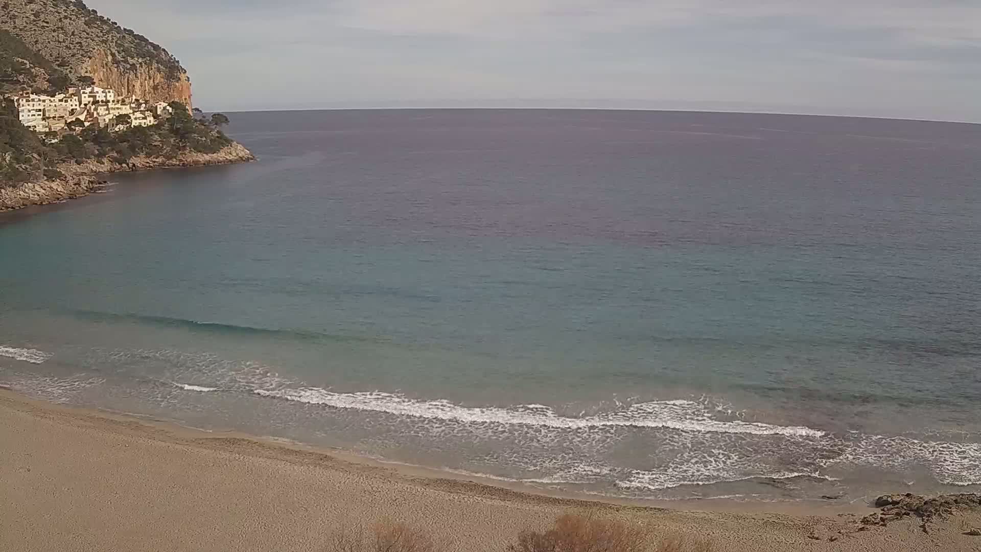 A sandy beach with small waves laps the shore under a cloudy sky, next to a rocky cove with buildings on a hillside.