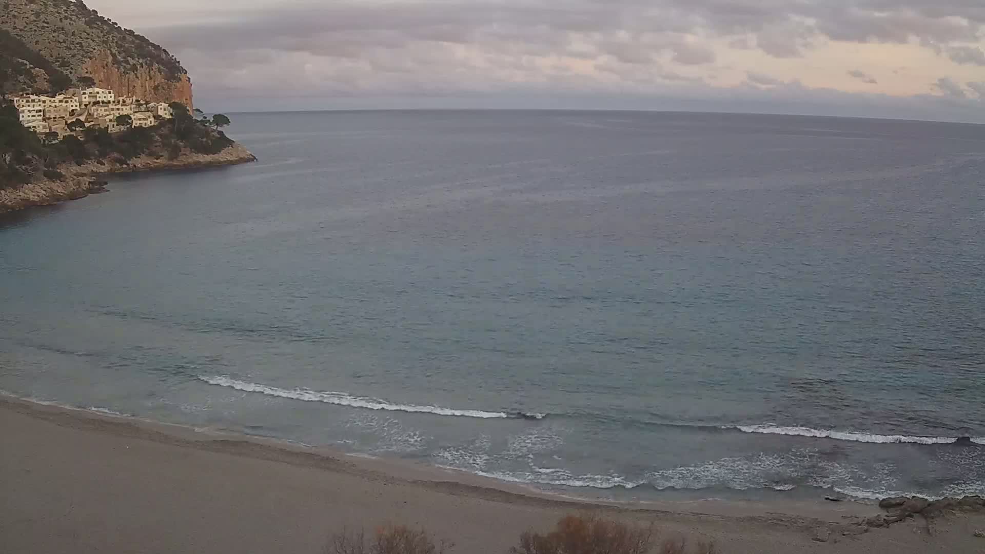 A sandy beach with small waves meets a calm sea, backed by a rocky cliff with white buildings under a cloudy sky.