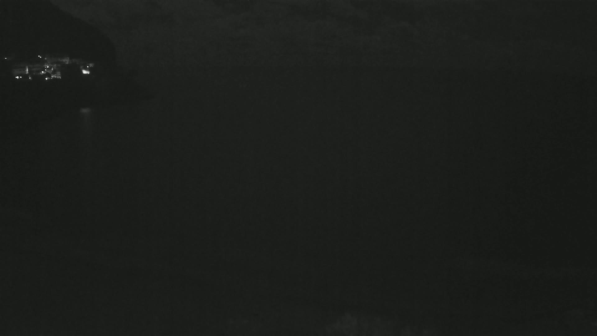 A dark, nighttime scene shows a body of water reflecting faint lights from a distant, mountainous shoreline under a cloudy sky.