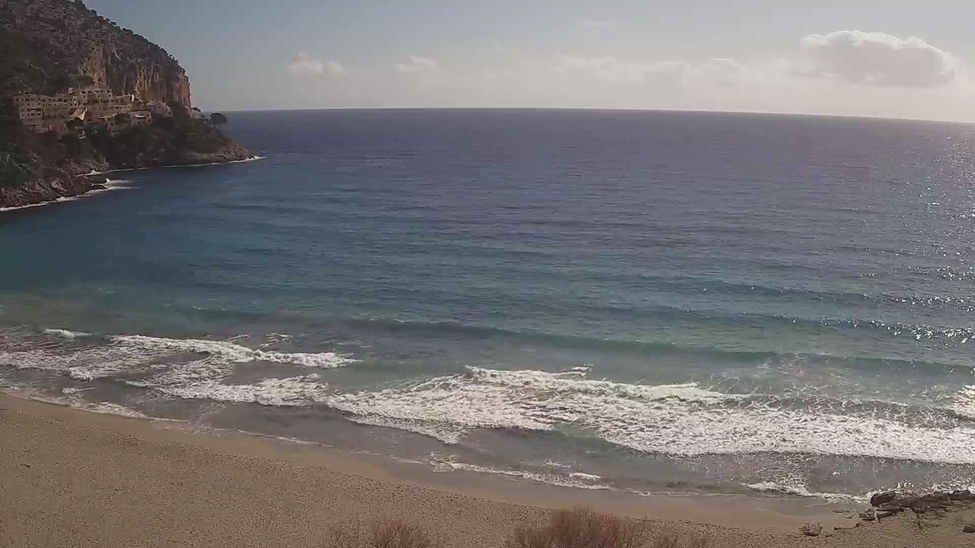 A sandy beach meets a calm, turquoise ocean with small waves lapping the shore, backed by a rocky cliff with buildings on its side under a partly cloudy sky.