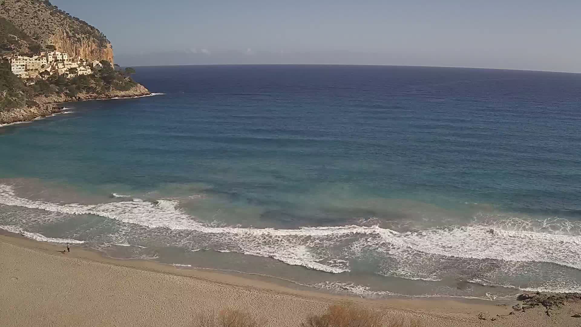A sandy beach with gentle waves laps the shore, under a clear sky, next to a rocky cliff with buildings on top.