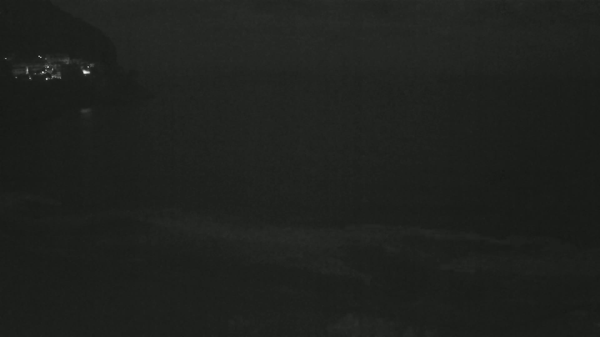 A dark, nighttime coastal scene shows a dimly lit hillside and dark water with subtle waves.