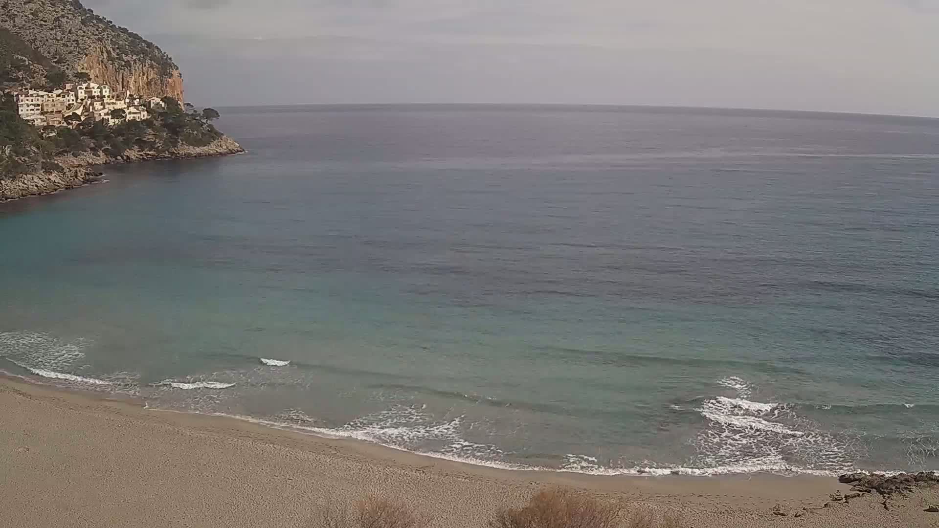 A sandy beach curves gently into a calm, teal sea lapping against the shore, under an overcast sky, backed by a rocky cliff with buildings perched upon it.