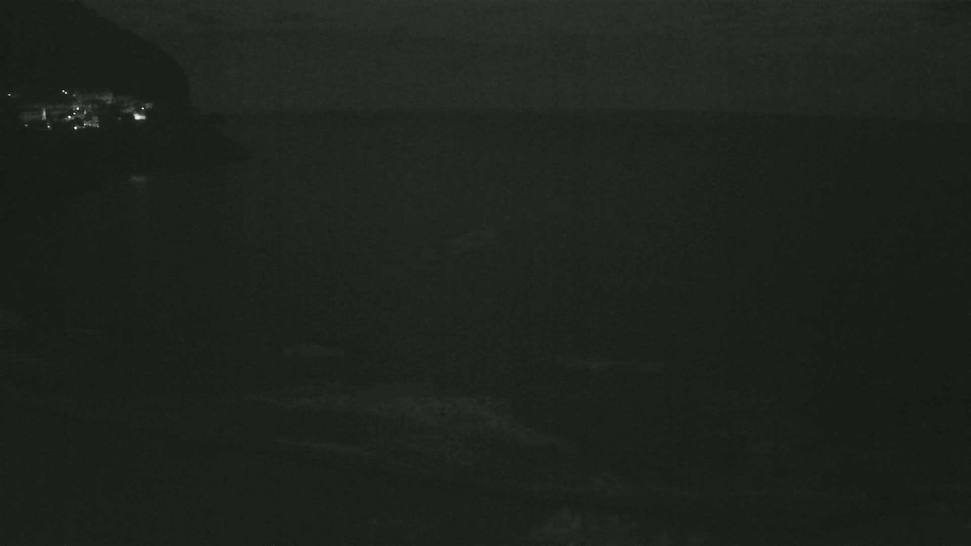 A dark, nighttime scene shows a body of water with a dimly lit shoreline and hillside in the distance.