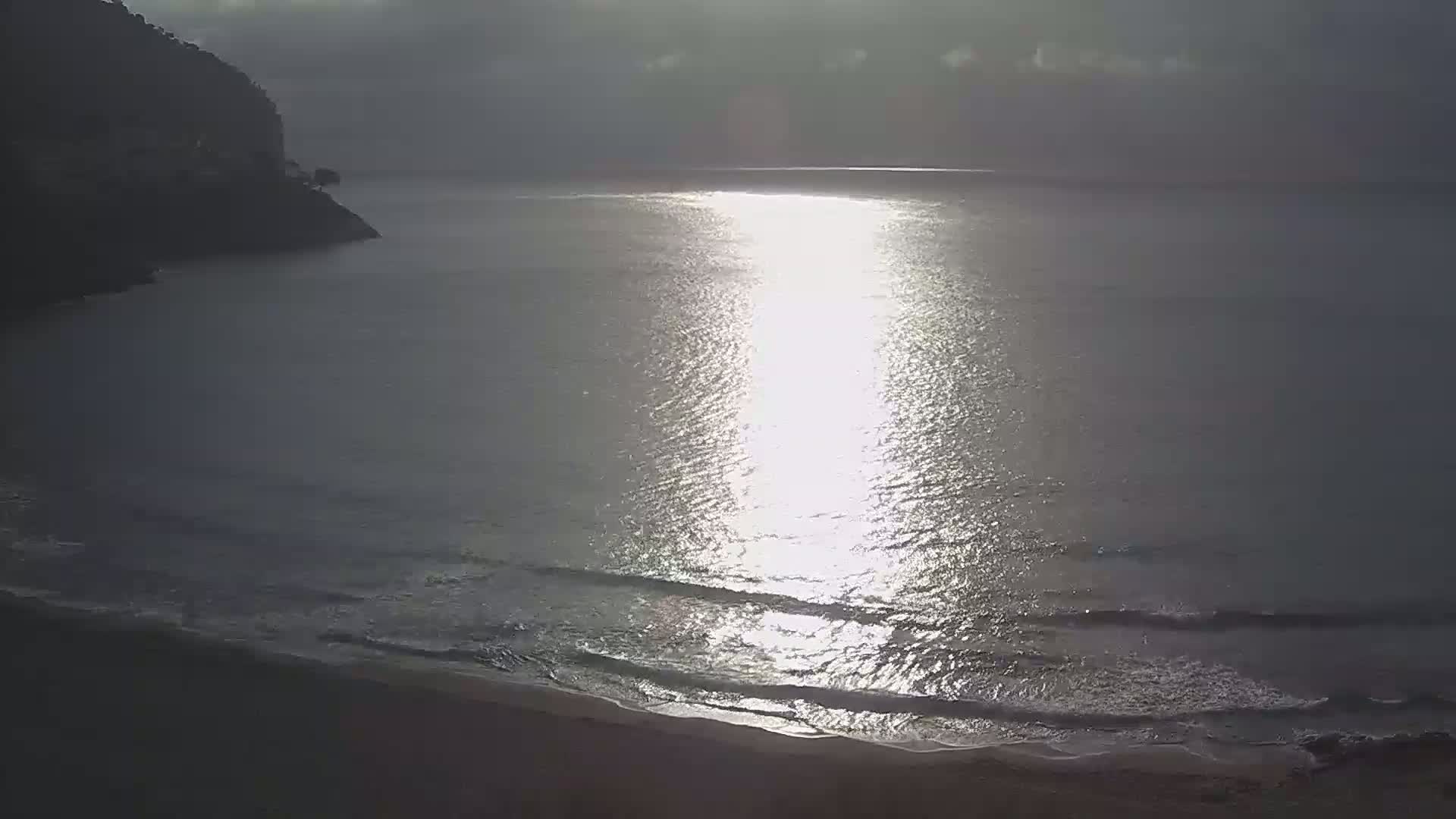 A tranquil beach scene shows gentle waves lapping a sandy shore under a partly cloudy sky, with the sun reflecting brightly on the calm water.