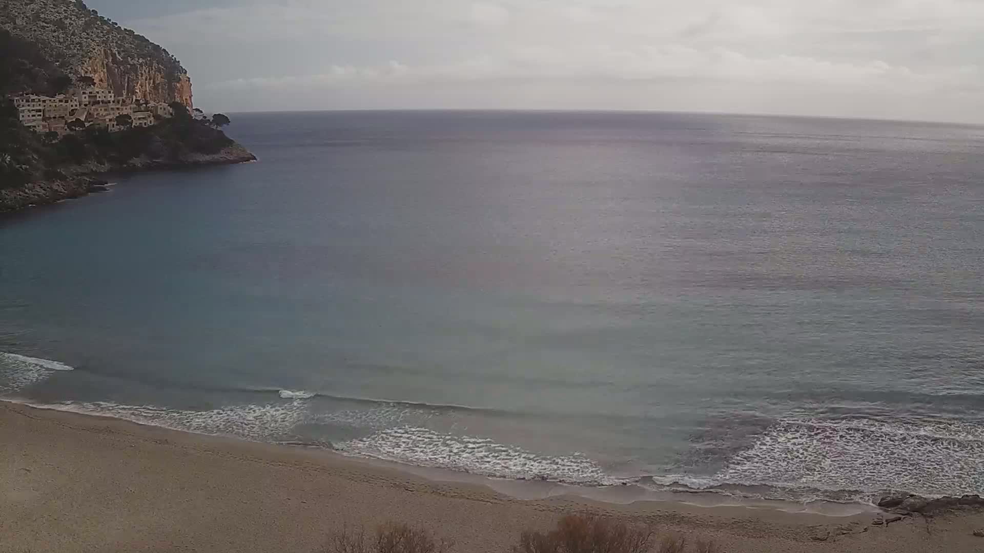A sandy beach with gentle waves laps against the shore under an overcast sky, nestled beside a rocky cliff with buildings perched on top.