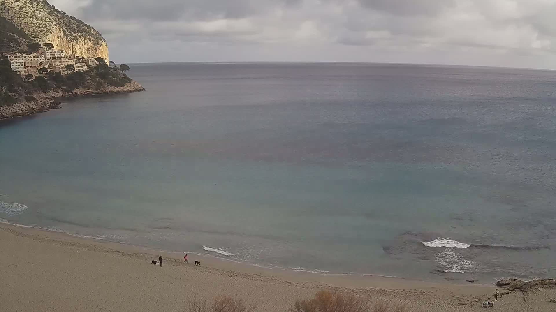 A sandy beach curves into a calm, gray-blue sea under a cloudy sky, with a few people walking their dogs along the shore and a rocky cliff with buildings visible in the background.