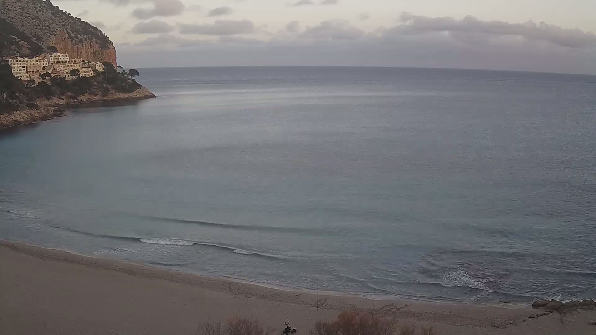 A sandy beach curves into a calm, gray-blue sea under a cloudy sky, backed by a rocky cliff with buildings on top.