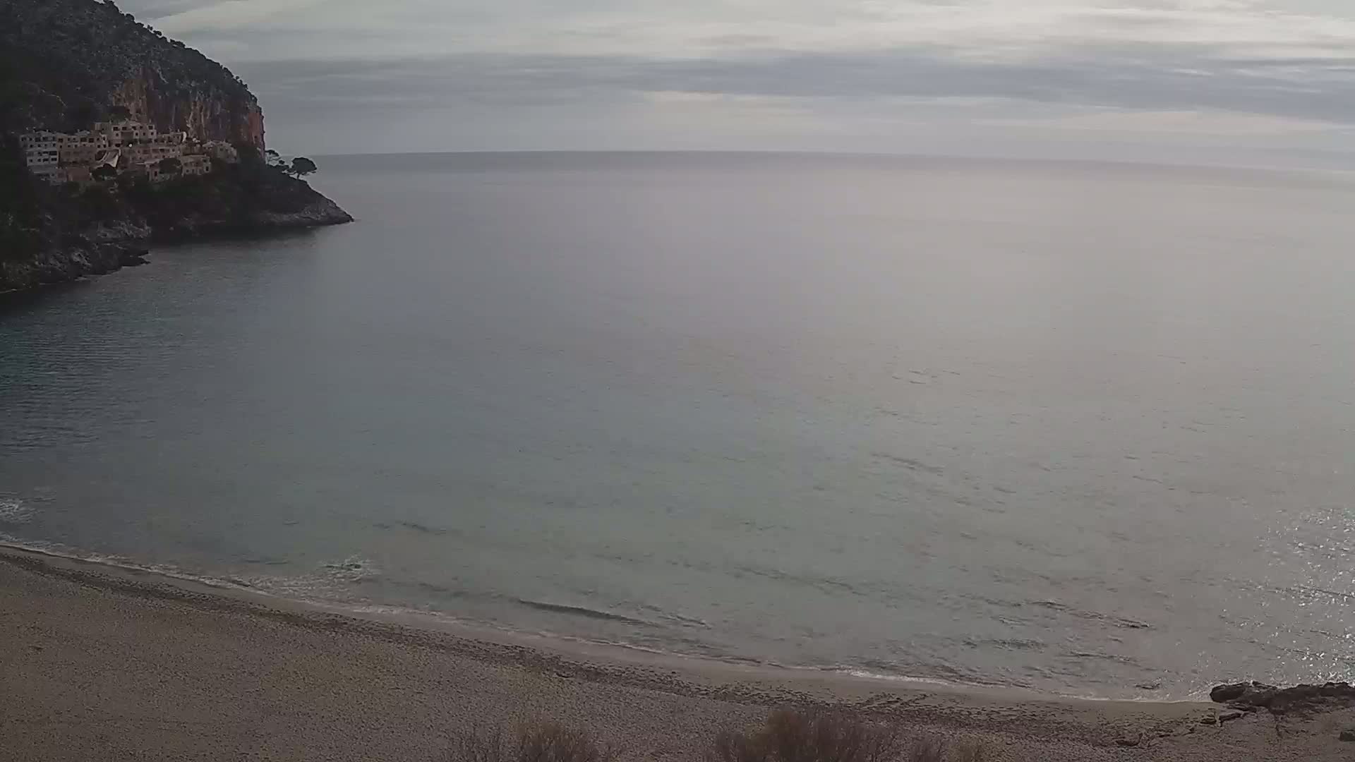 A sandy beach curves into a calm, gray-toned sea under a cloudy sky, with a rocky cliff and buildings visible in the background.