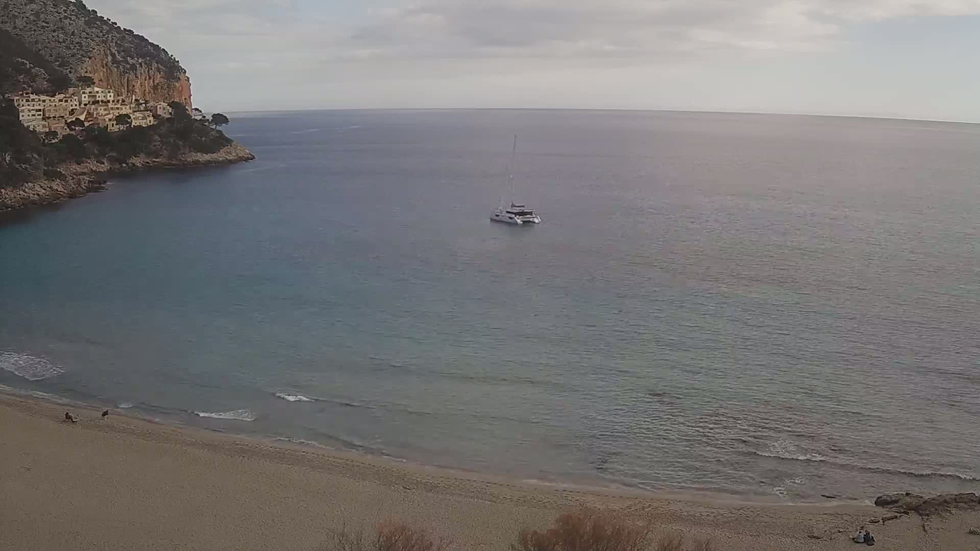 A catamaran floats in a calm sea near a sandy beach and a rocky cliff with buildings on top under an overcast sky.
