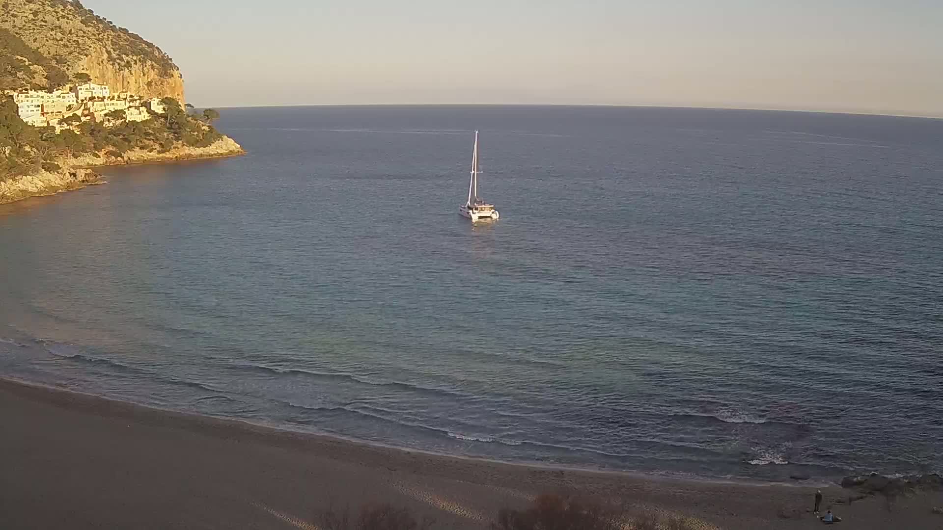 A sailboat sits at anchor in calm sea waters near a sandy beach and a rocky cliff with buildings on top under a clear, light sky.