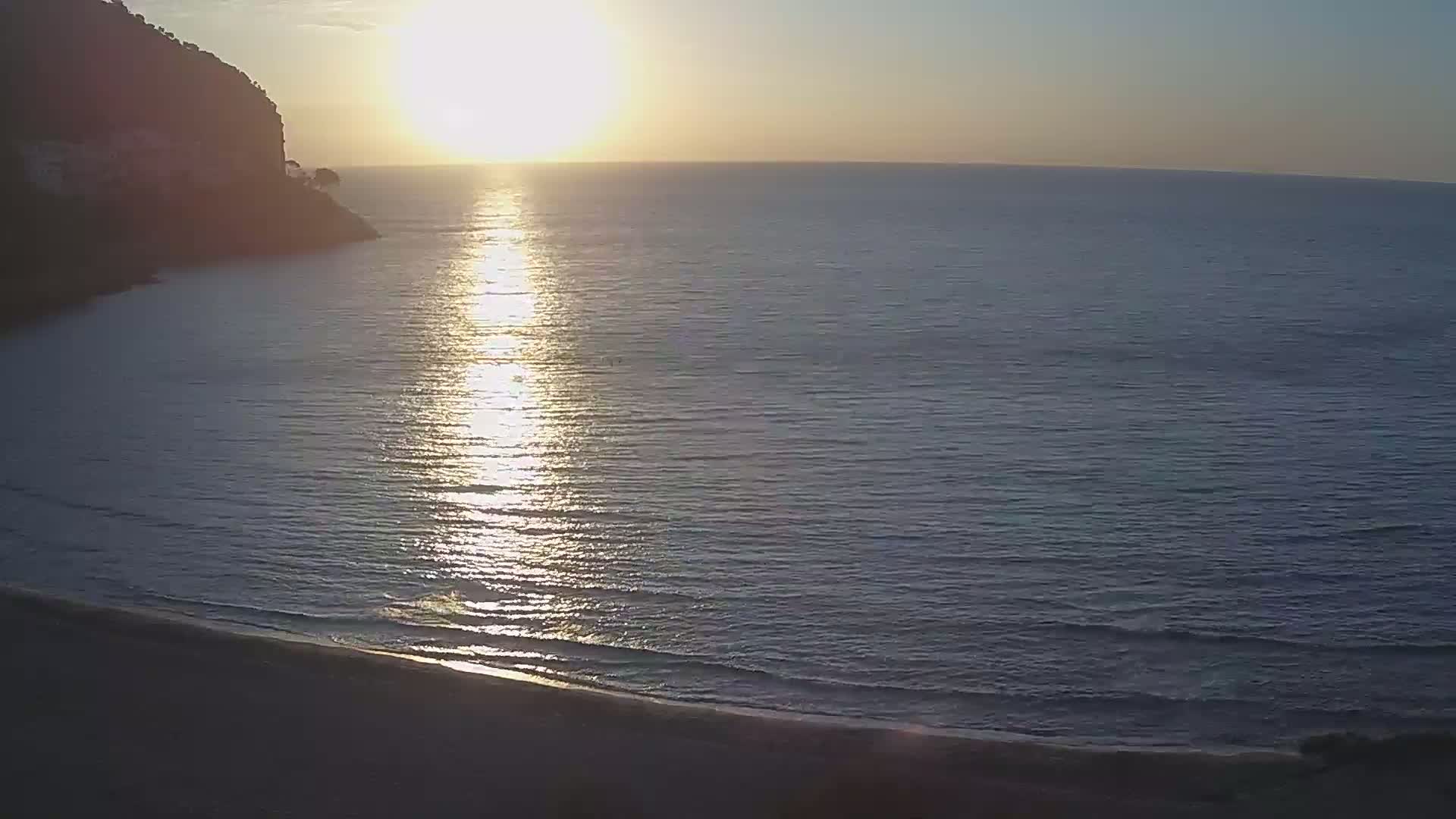 A tranquil beach scene at sunset shows the sun reflecting on calm ocean water lapping a sandy shore next to a dark cliff.