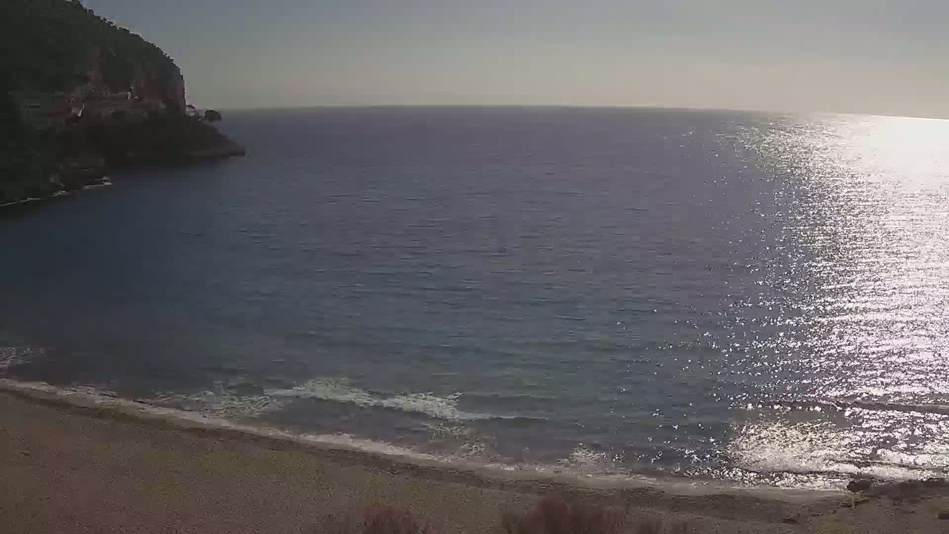 A rocky cliff overlooks a calm, sunlit sea with a small, sandy beach.