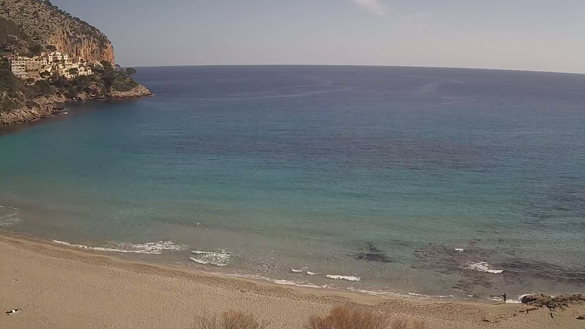 A sandy beach meets clear, turquoise water under a sunny sky, backed by a rocky cliff with buildings on top.