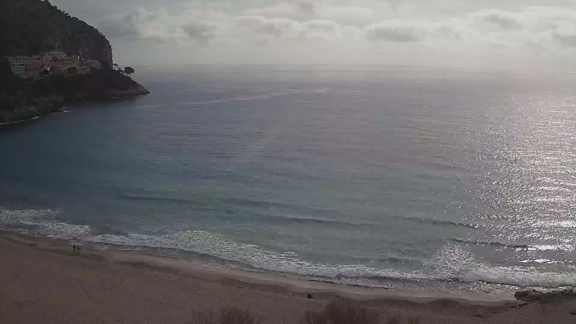 A sandy beach with gentle waves laps the shore under a partly cloudy sky, adjacent to a rocky cliff with buildings on its peak.