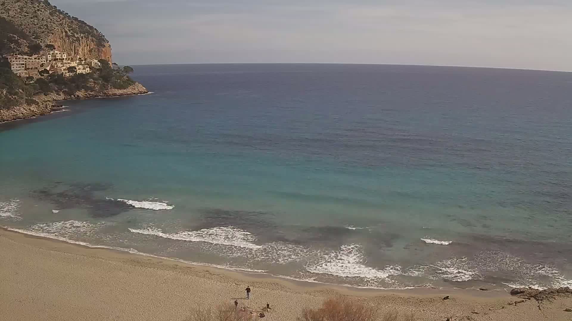 A sandy beach with gentle waves laps at the shore of a turquoise sea, backed by a rocky cliff with buildings on top, under a mostly clear sky.