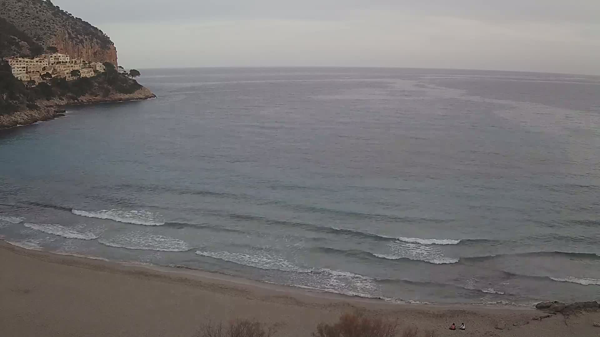 A sandy beach with small waves laps the shore under an overcast sky, beside a cove and a cliff with buildings on top.