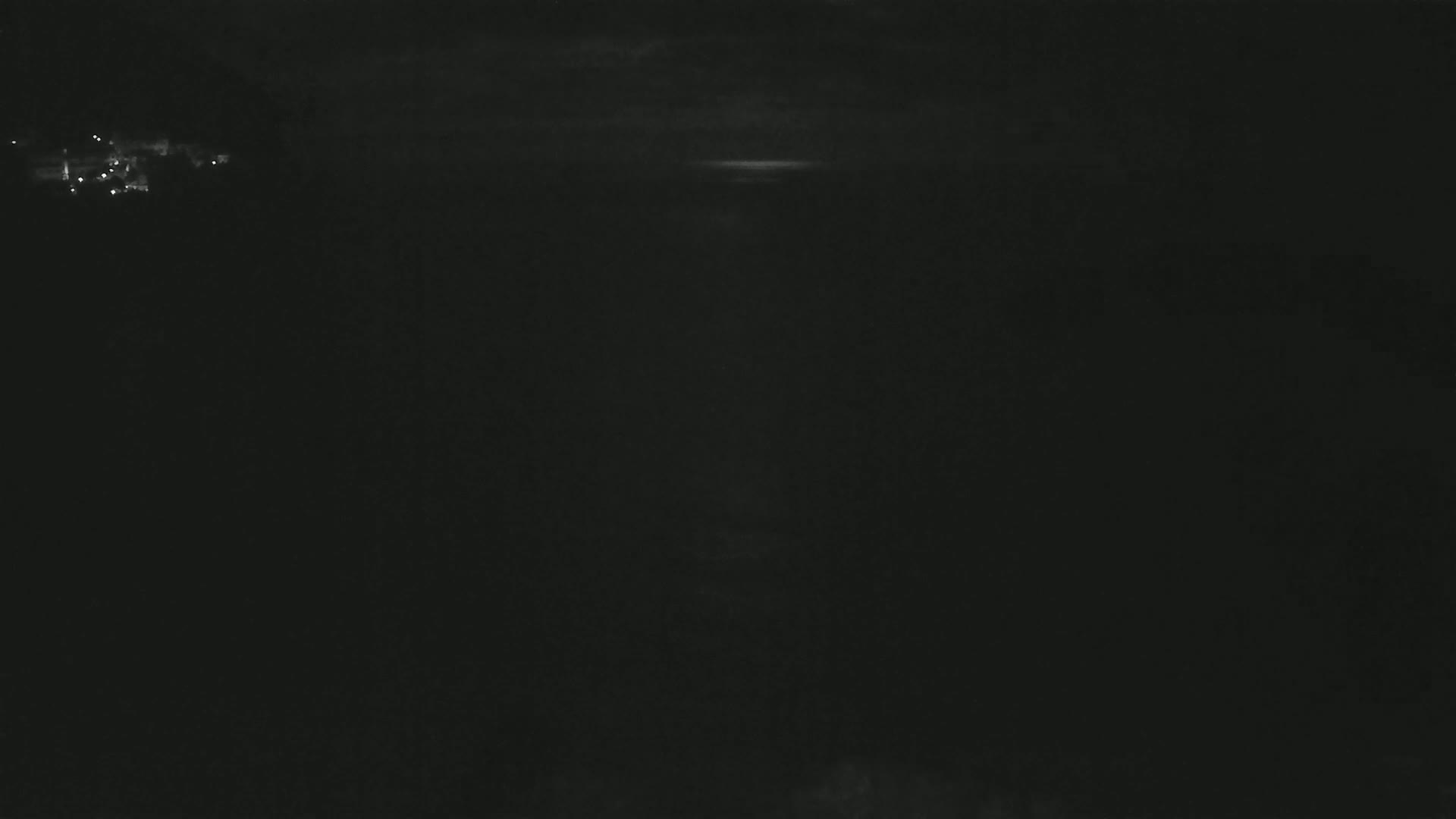 A dark, nighttime scene shows a faint glow on the horizon under a cloudy sky, with a few distant lights visible in the upper left.