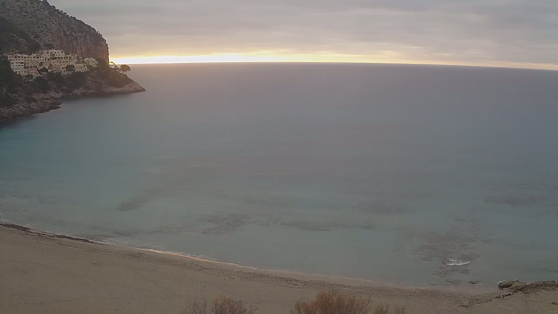 A sandy beach curves into a calm, teal sea at sunrise, bordered by a rocky cliff with buildings perched atop it under an overcast sky.