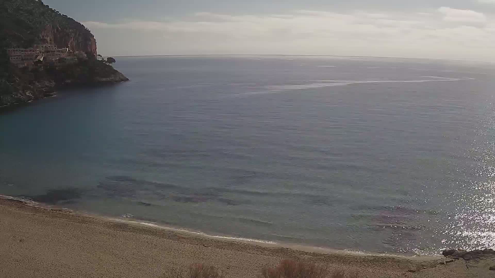 A sandy beach curves into a calm, blue sea, backed by a rocky cliff with buildings on its face under a mostly clear sky.