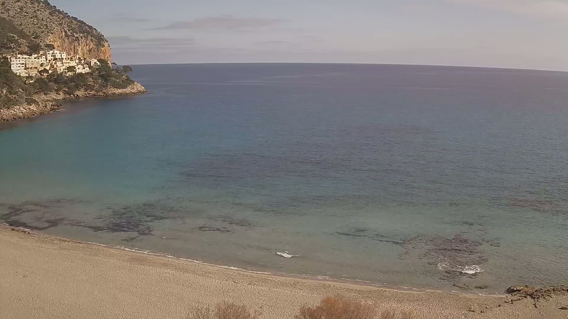 A sandy beach curves into a calm, clear turquoise ocean, bordered by a rocky cliff with buildings on top under a mostly clear sky.
