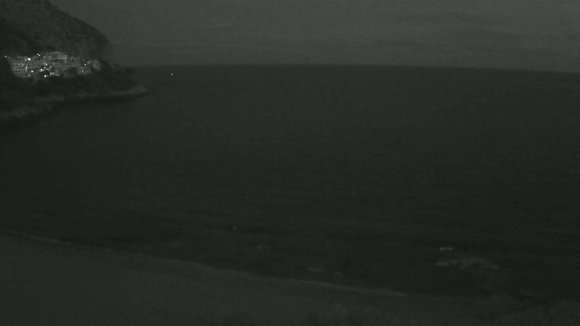 A dark, nighttime scene shows a rocky cliff with lights visible on the hillside overlooking a calm, dark sea.