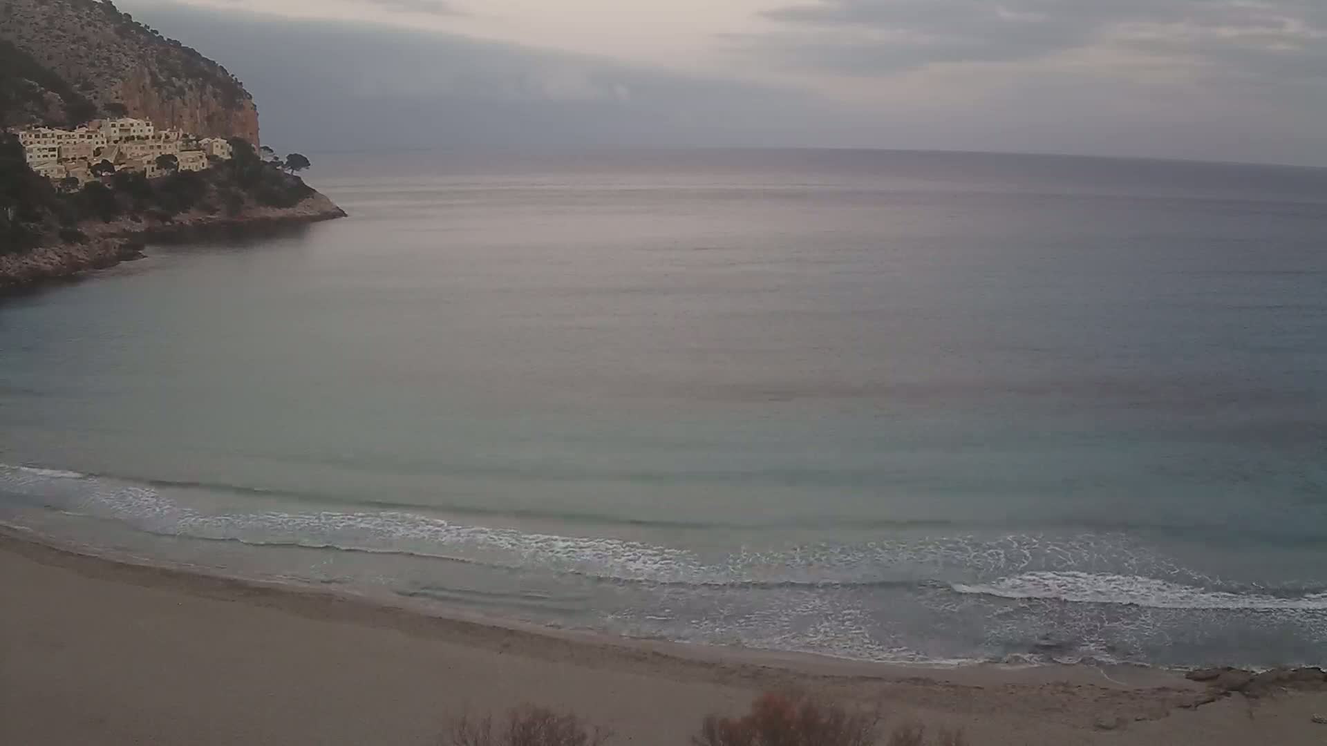 A sandy beach with gentle waves laps the shore under an overcast sky, nestled in a cove next to a rocky cliff with buildings on top.