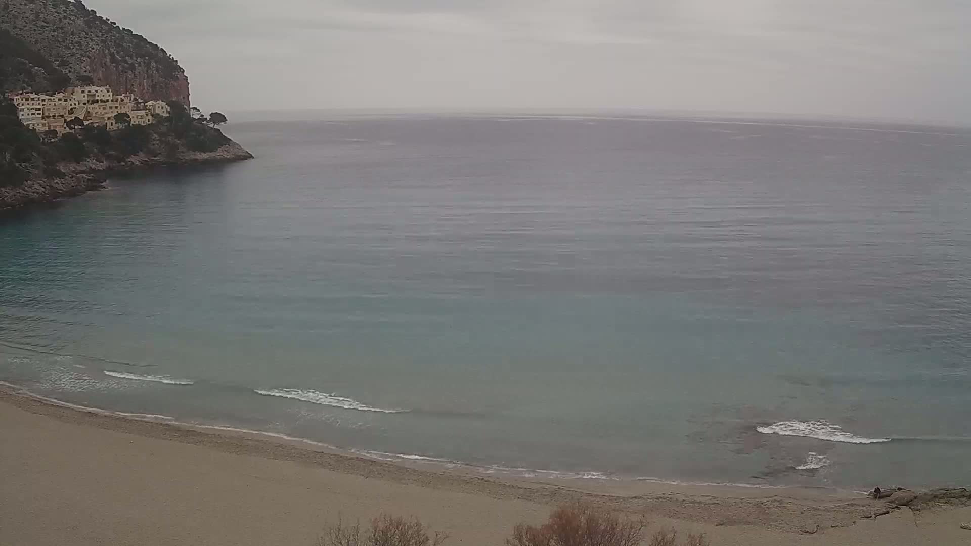 A sandy beach curves into a calm, gray-toned sea lapping against the shore, nestled beneath a cliffside with buildings.
