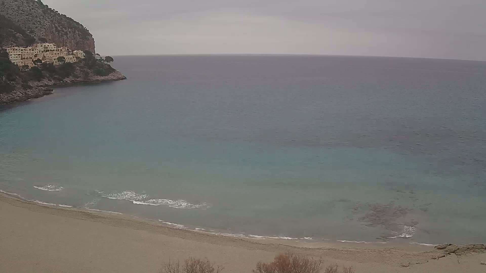 A sandy beach curves into a calm, teal sea under a cloudy sky, with buildings perched atop a rocky cliff in the background.