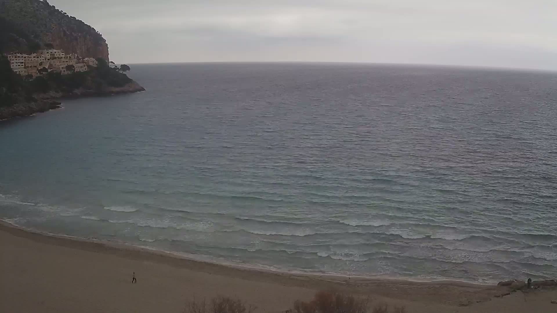 A sandy beach with gentle waves laps the shore under an overcast sky, with a rocky cliff and buildings visible in the background.