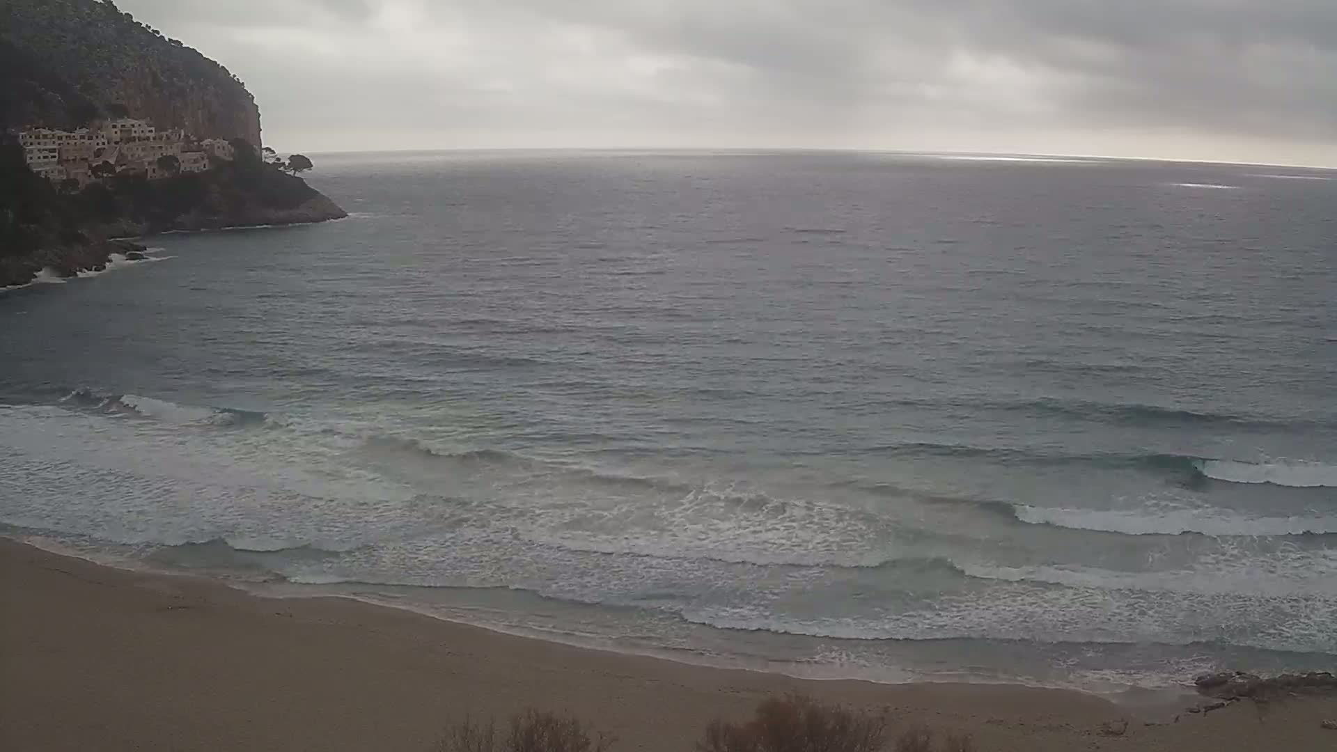 A sandy beach meets a calm sea with small waves under an overcast sky, with a cliff and buildings visible in the distance.