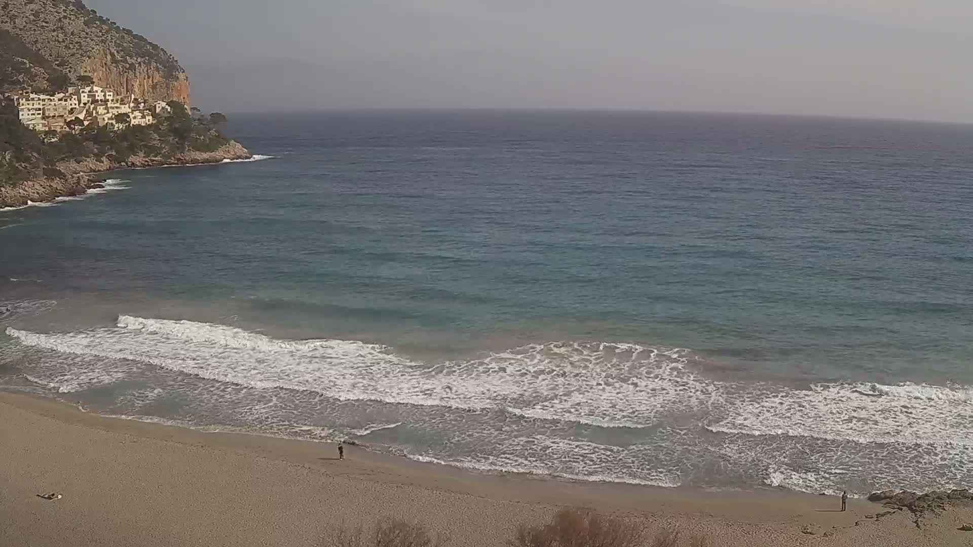A sandy beach with gentle waves laps the shore under a hazy sky, near a rocky cliff with buildings on top.