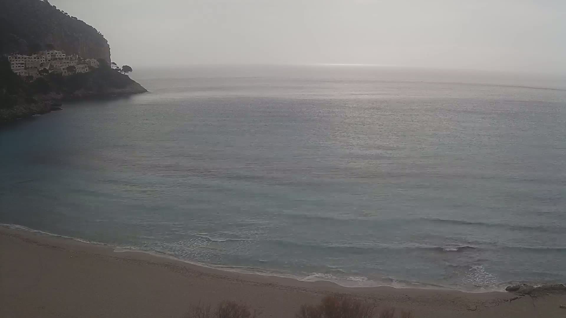 A sandy beach meets a calm, grayish-blue sea under an overcast sky, with a cliff and buildings visible in the distance.