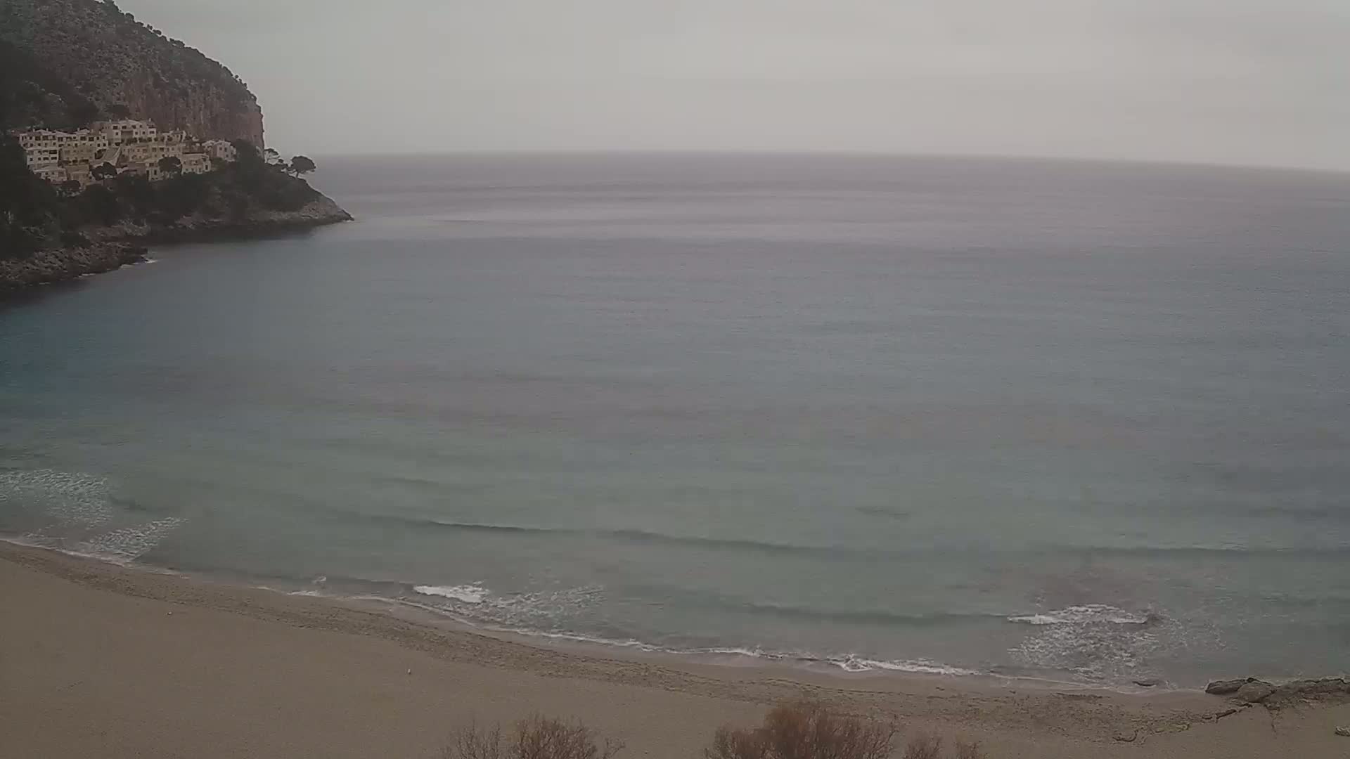 A sandy beach curves into a calm, grey-toned sea under an overcast sky, with a rocky cliff and buildings visible in the background.