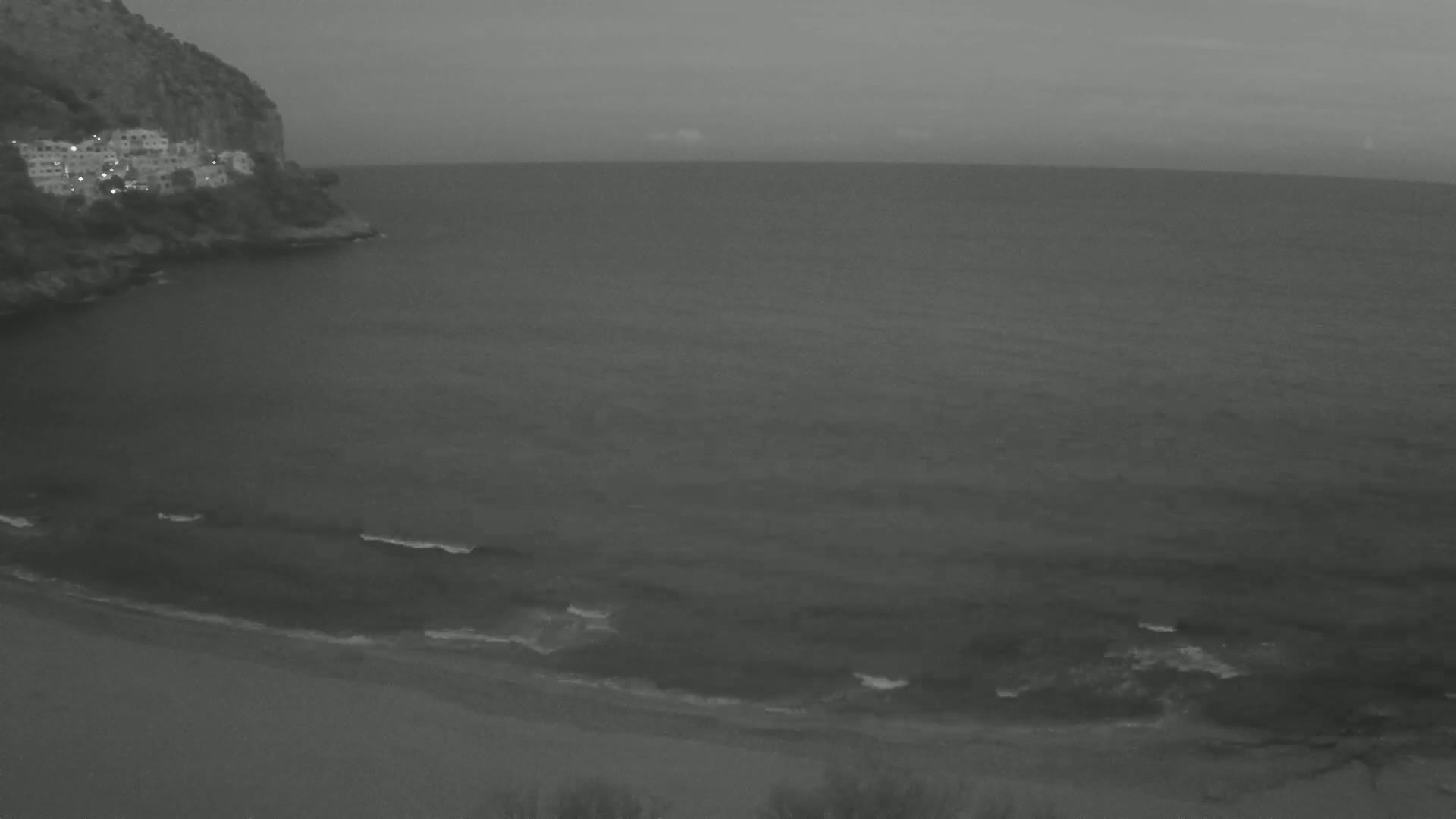 A grayscale image shows a rocky coastline with buildings nestled into the hillside, overlooking a calm sea with small waves gently lapping a sandy shore under an overcast sky.