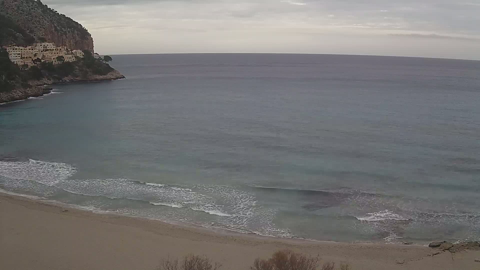 A sandy beach with small waves laps against the shore, under an overcast sky, next to a rocky cliff with buildings on top.