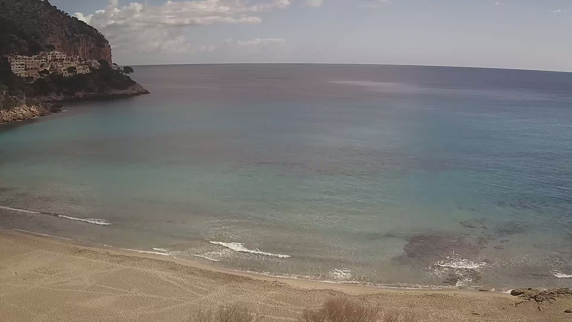 A sandy beach curves into a calm, teal sea under a partly cloudy sky, backed by a rocky cliff with buildings on top.