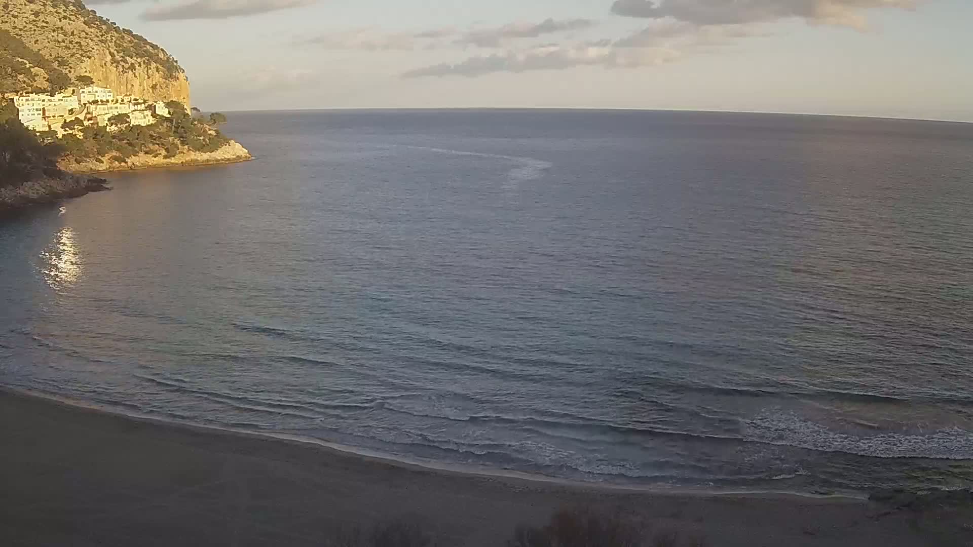A sandy beach meets a calm, gray-blue sea under a partly cloudy sky, with a rocky cliff and buildings visible in the background.
