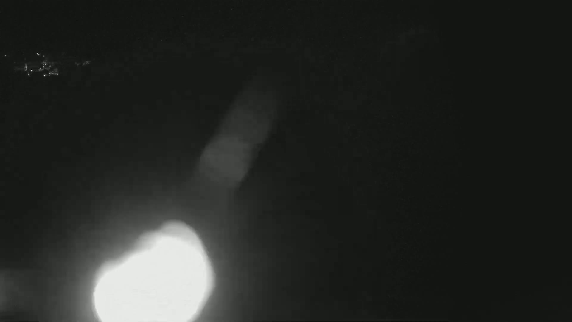 A bright light source, possibly headlights, is visible in a dark, nighttime setting with faint light sources in the distance.