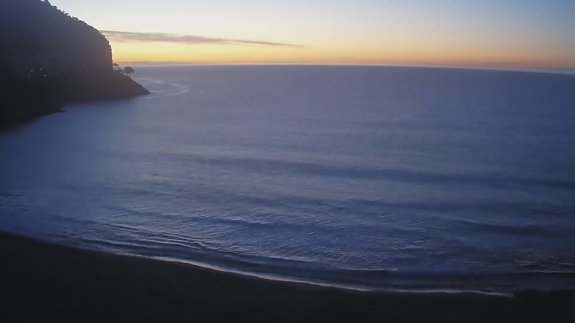 A dark, rocky shoreline curves into a calm ocean under a dusky, sunrise sky.