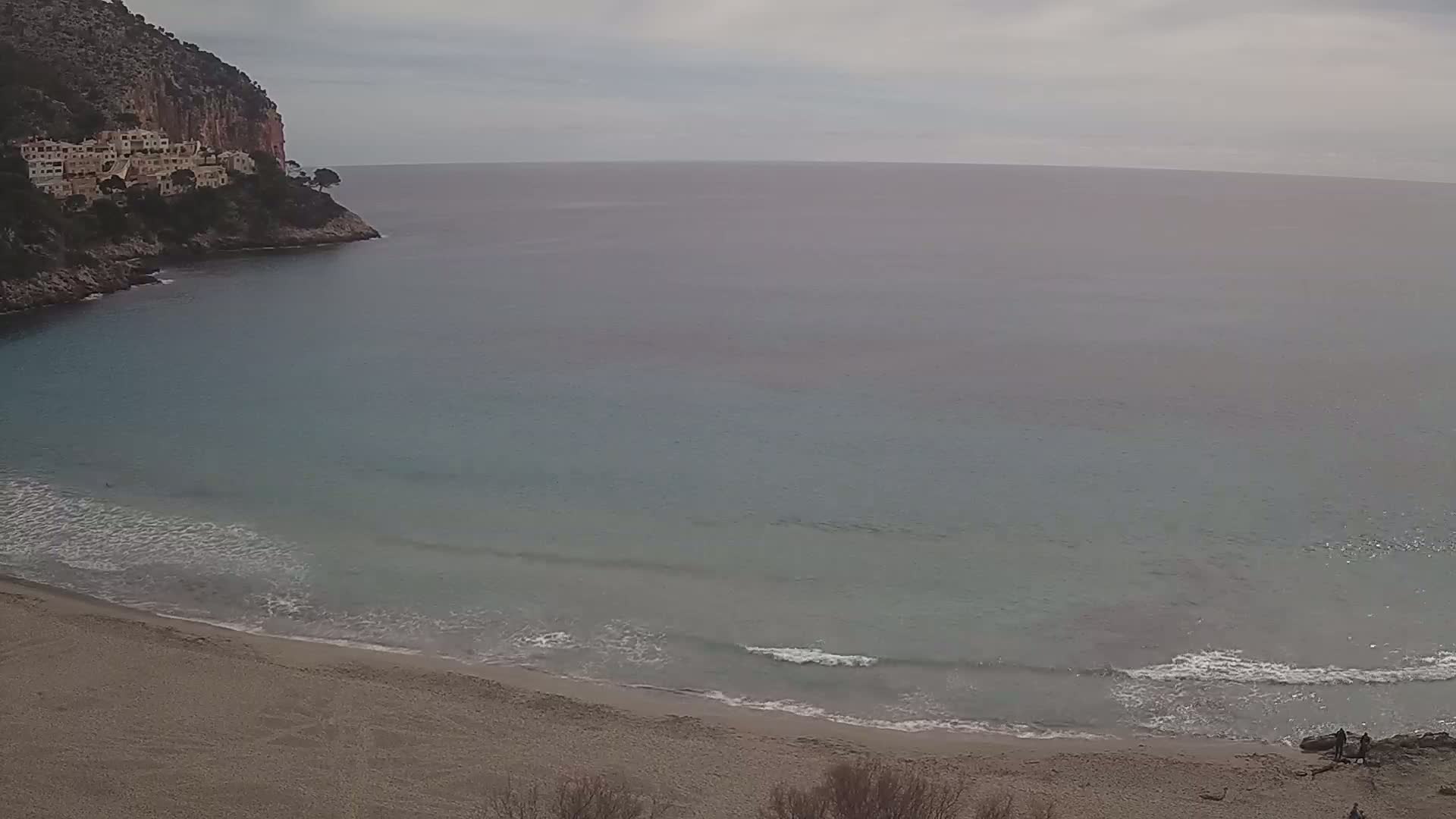 A sandy beach with small waves laps the shore of a calm, teal ocean, nestled below a cliffside with buildings on a cloudy day.
