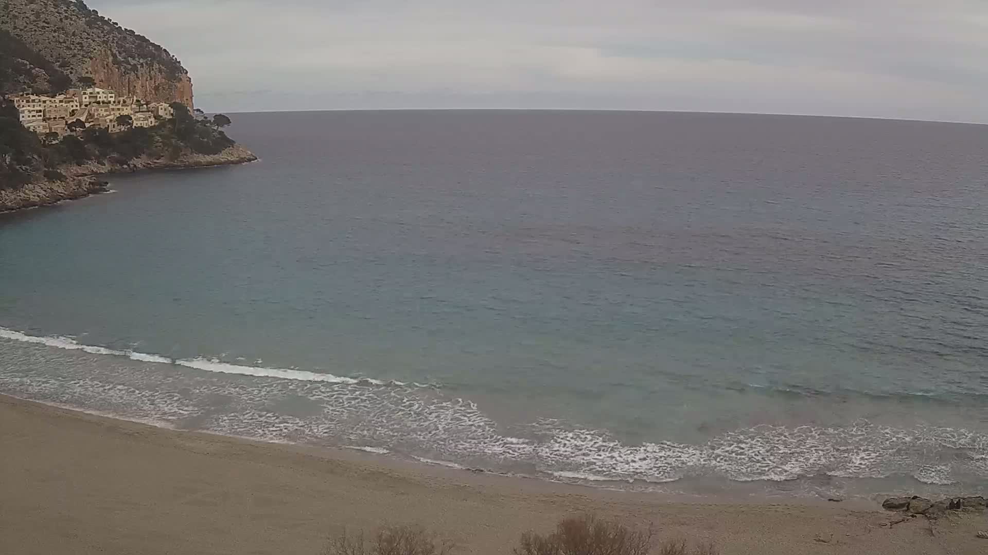 A sandy beach with gentle waves laps the shore, under an overcast sky, nestled in a cove next to a rocky cliff with buildings.