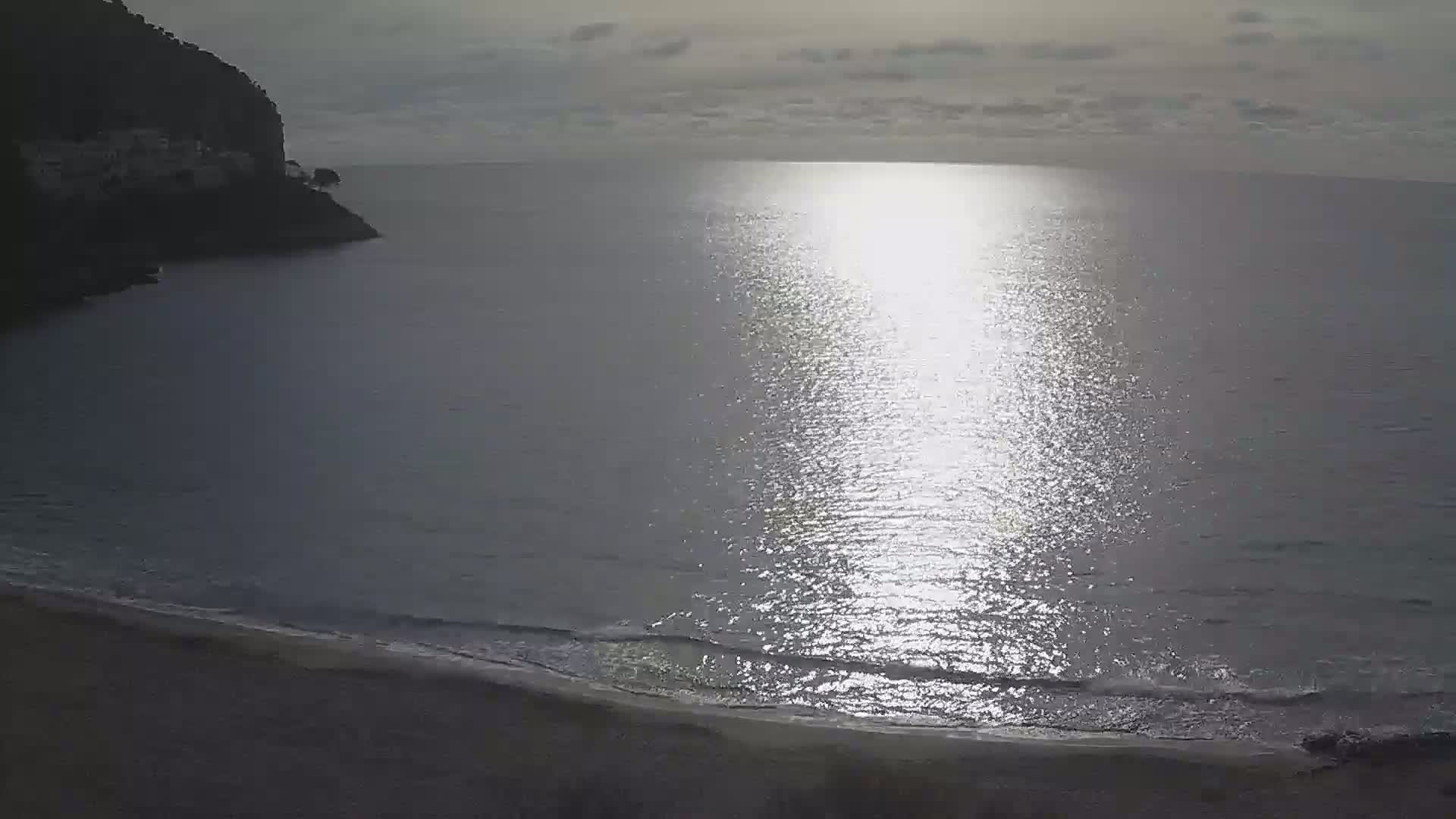 A dark, rocky cliff overlooks a calm, sunlit sea with a sandy beach.