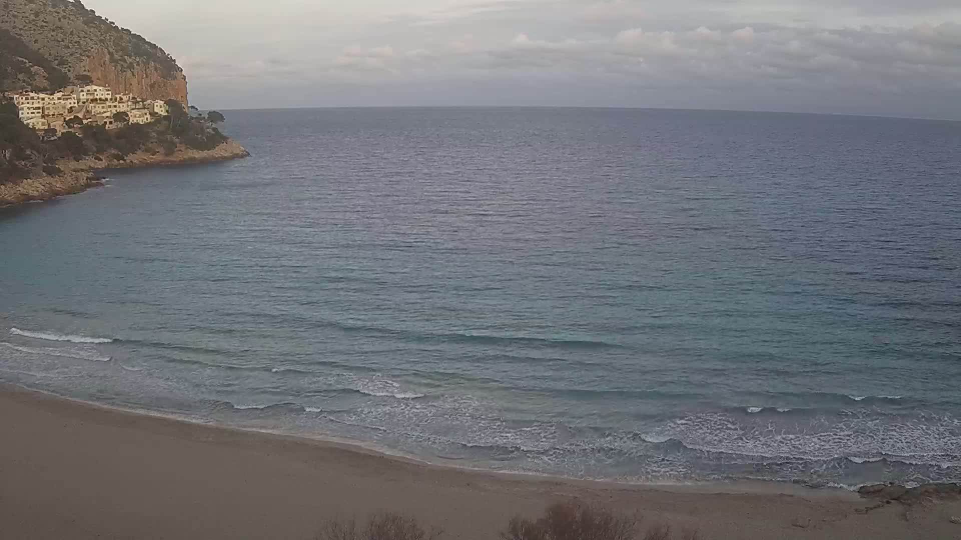 A sandy beach with gentle waves laps the shore under a cloudy sky, next to a rocky cliff with buildings on top.