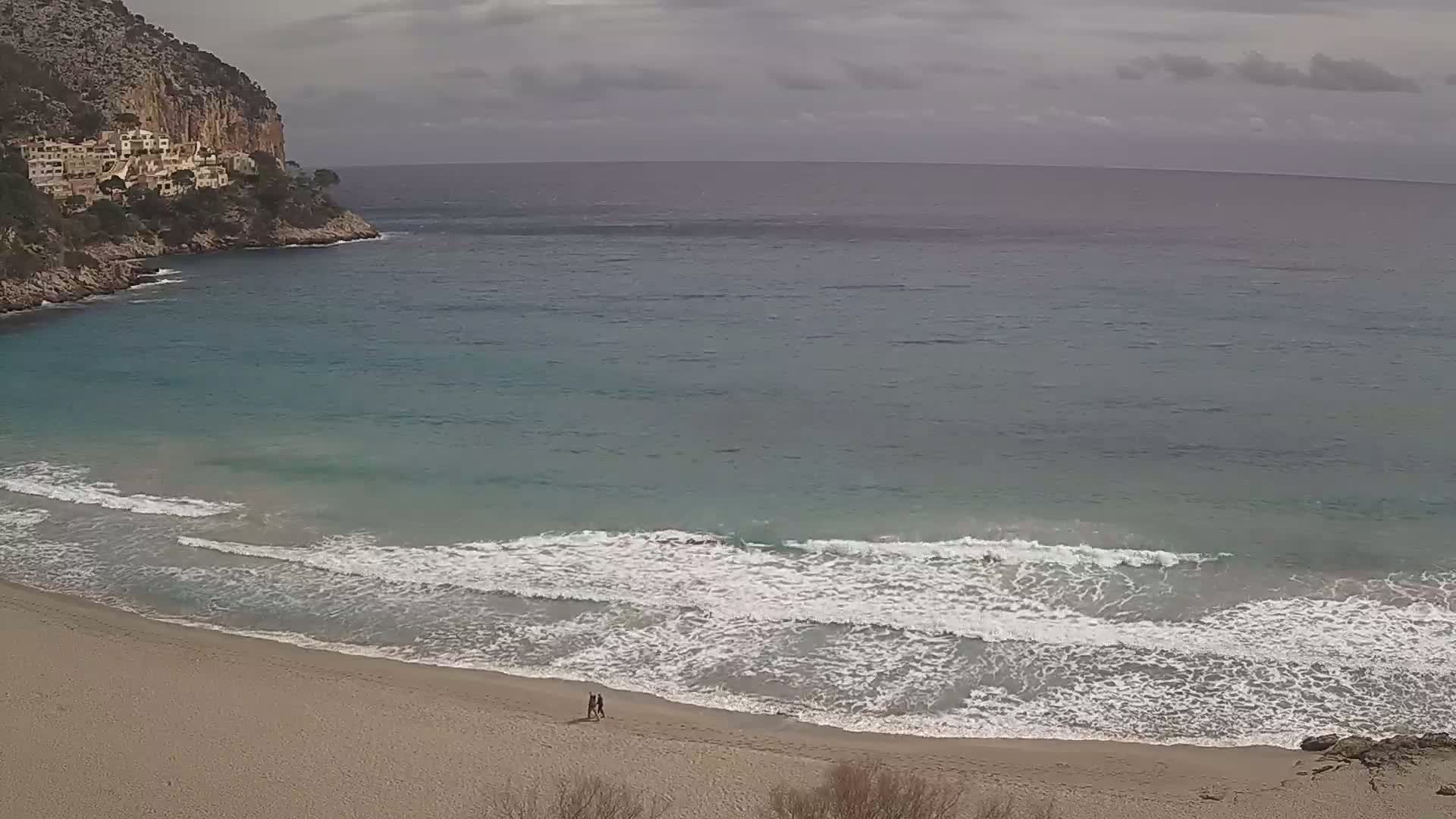 A sandy beach with small waves laps the shore under a cloudy sky, with a rocky cliff and buildings visible in the background.